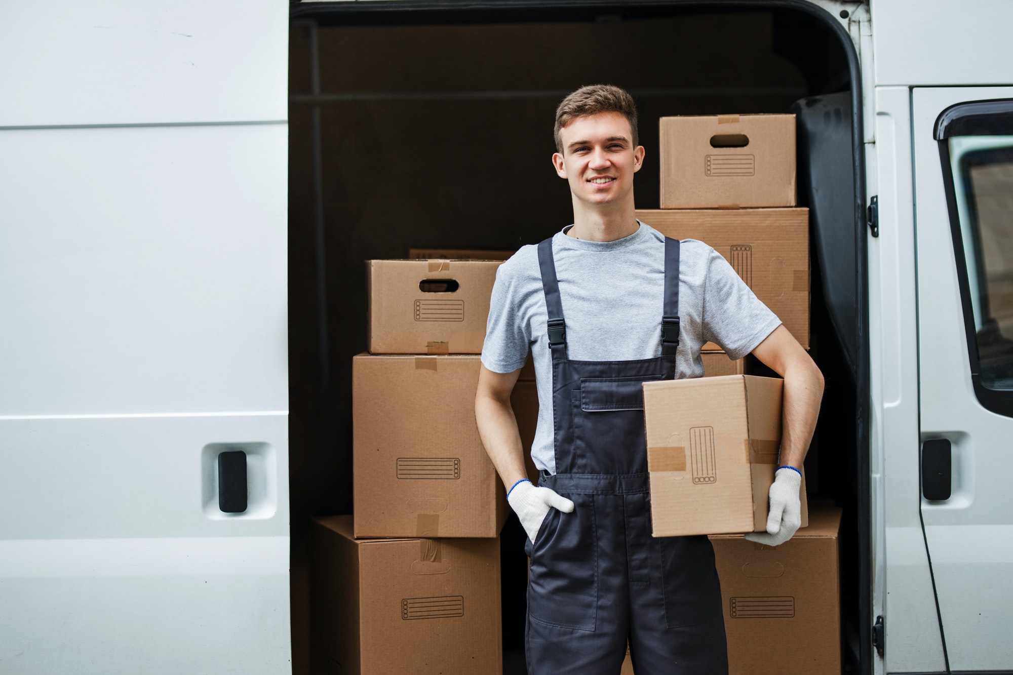 a-young-handsome-smiling-worker-wearing-uniform-is-standing-next-to-the-van-full-of-boxes-holding-a